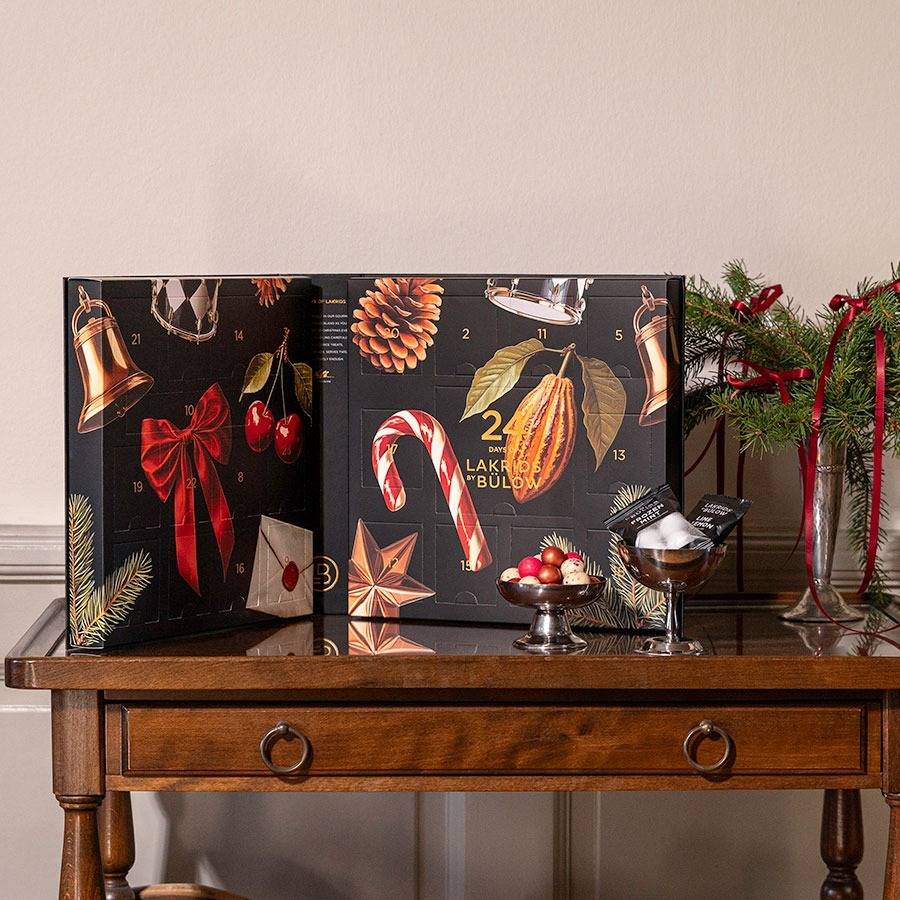 Chokladshopen Lakrids By Bülow adventskalender med svart och guldig juldesign på ett träbord med juldekorationer.